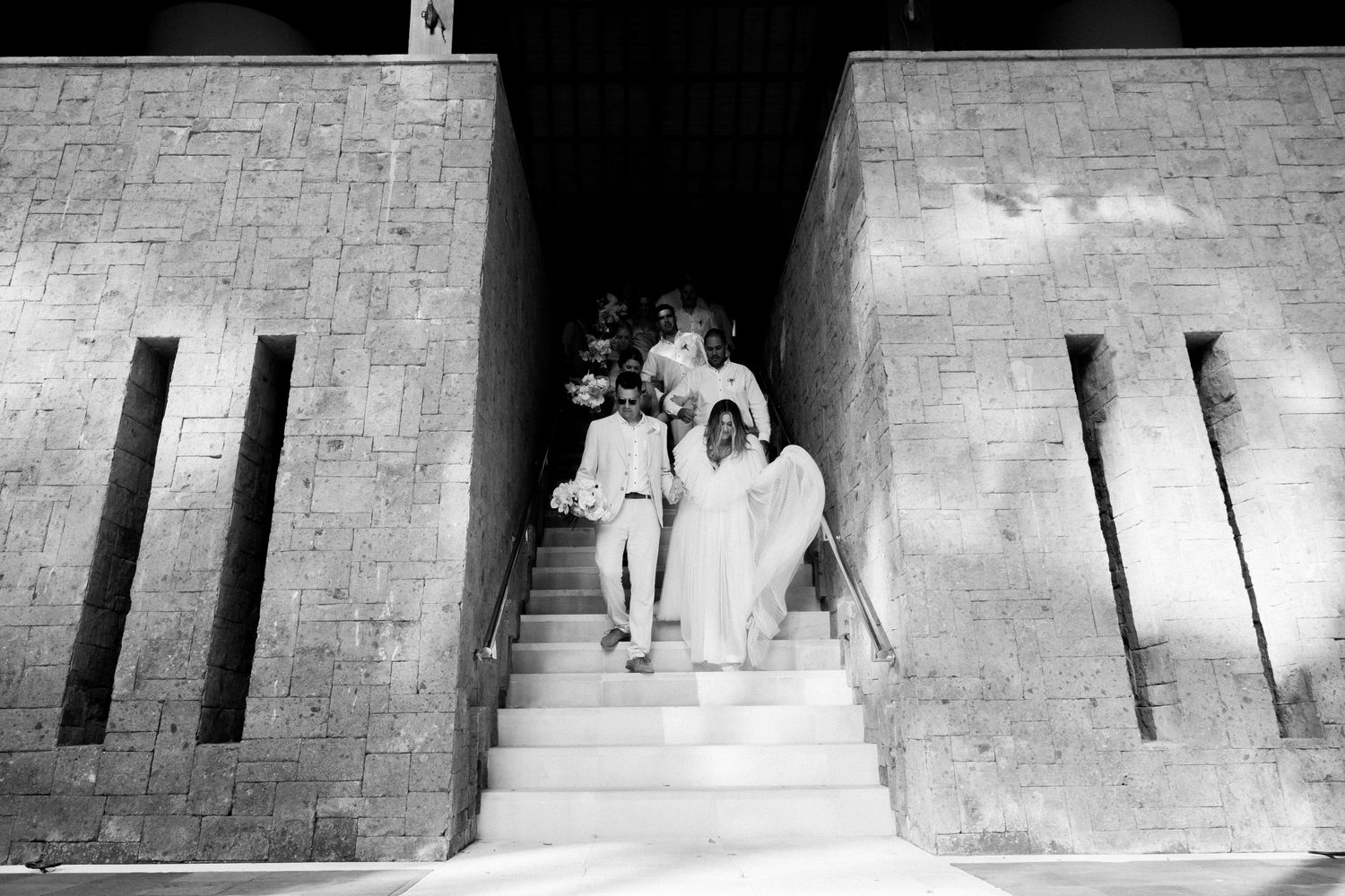 A black and white wedding scene shows a couple descending stone steps between angular architectural walls.