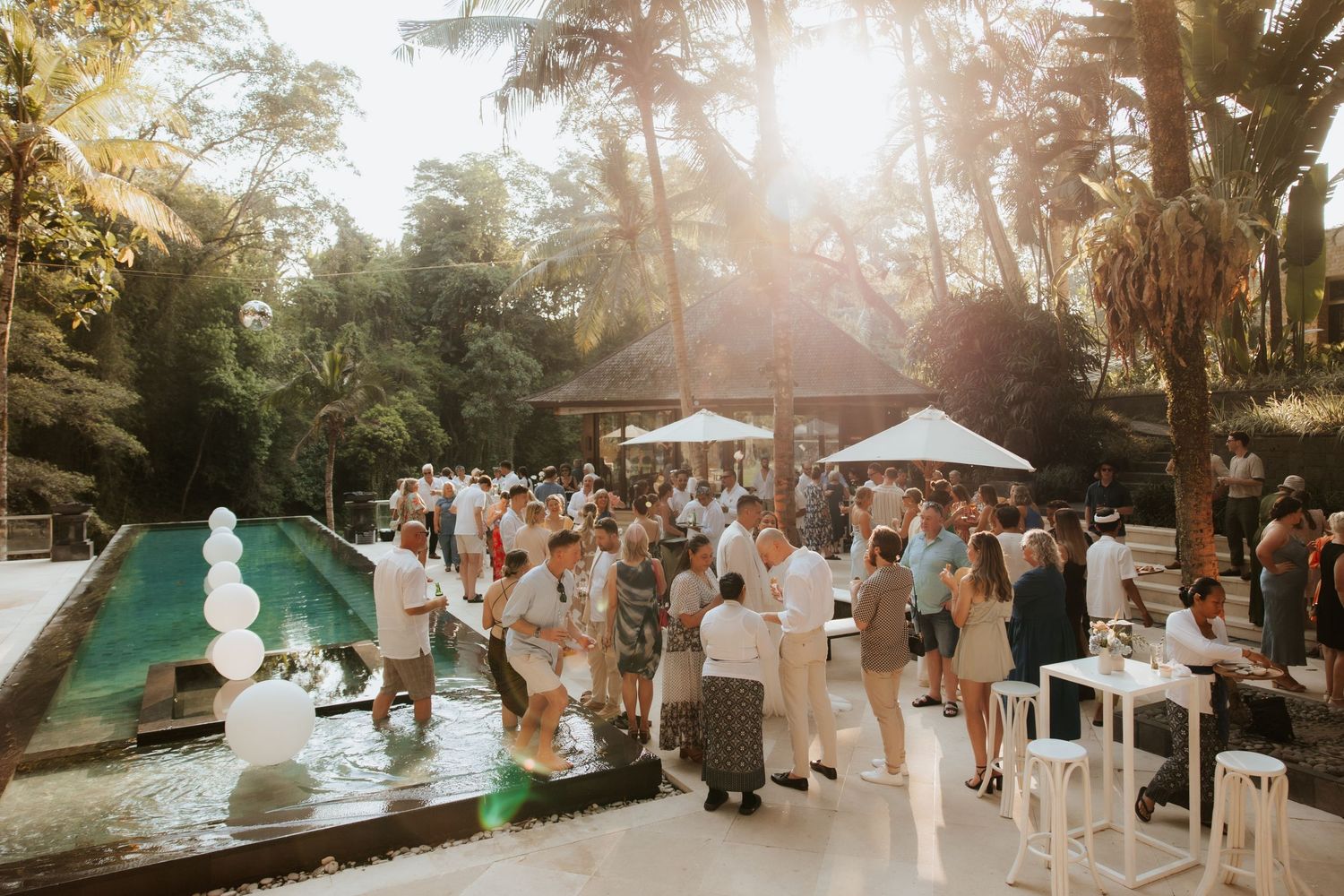 Outdoor wedding reception by a pool with guests mingling under white canopies as sunlight filters through tropical trees.