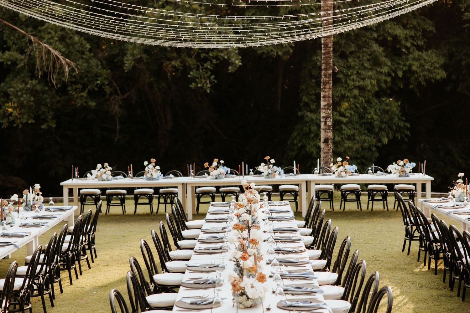 Long banquet tables set with white linens and decor under string lights at an outdoor reception venue.