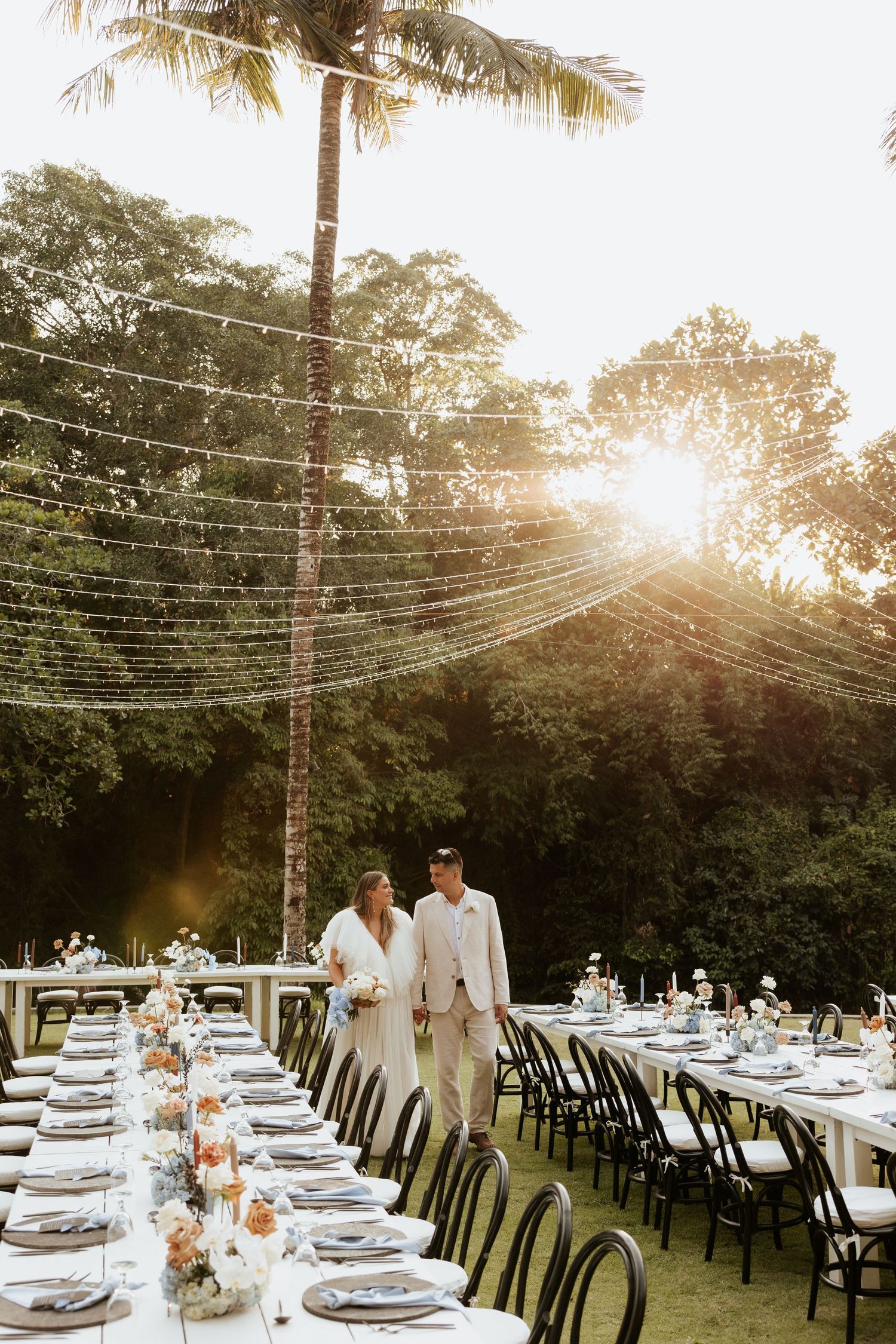 Wedding reception tables set under string lights and palm trees at sunset.