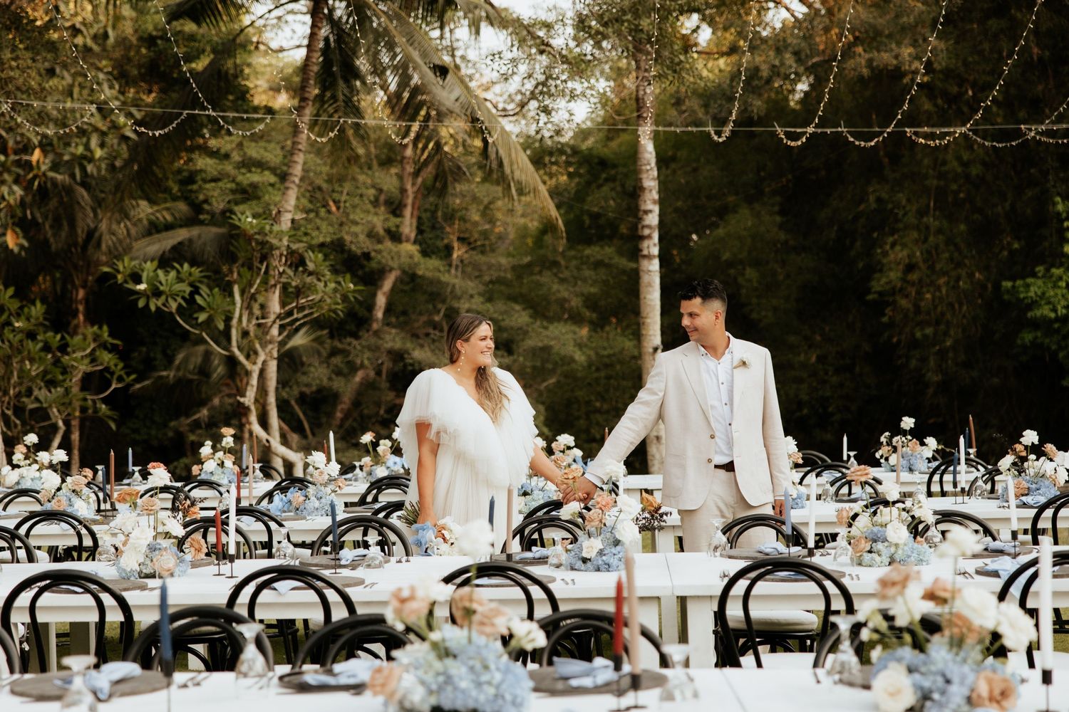Elegant outdoor wedding reception setup with bentwood chairs and blue floral centerpieces in a forest setting.
