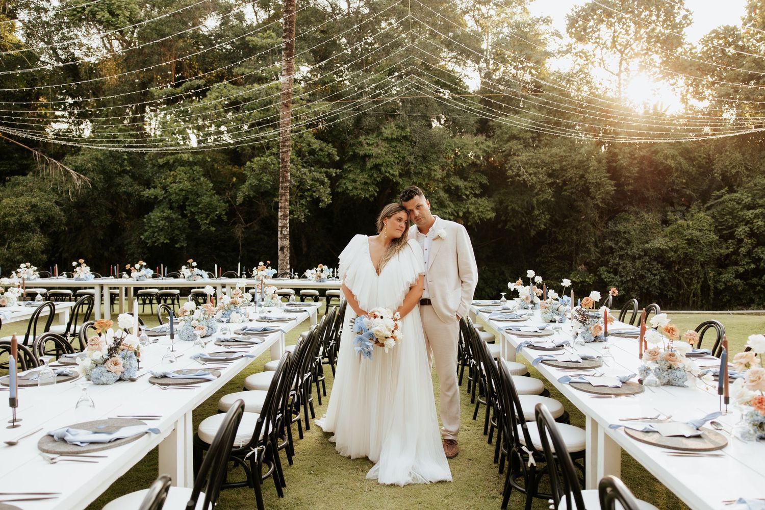 Elegant outdoor reception setup with long tables, string lights, and floral centerpieces in a garden setting at sunset.