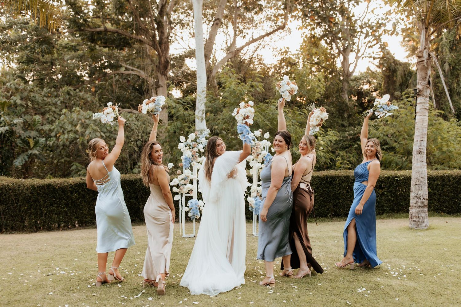 A joyful wedding party throws confetti in the air at golden hour in a lush garden setting with tall trees.
