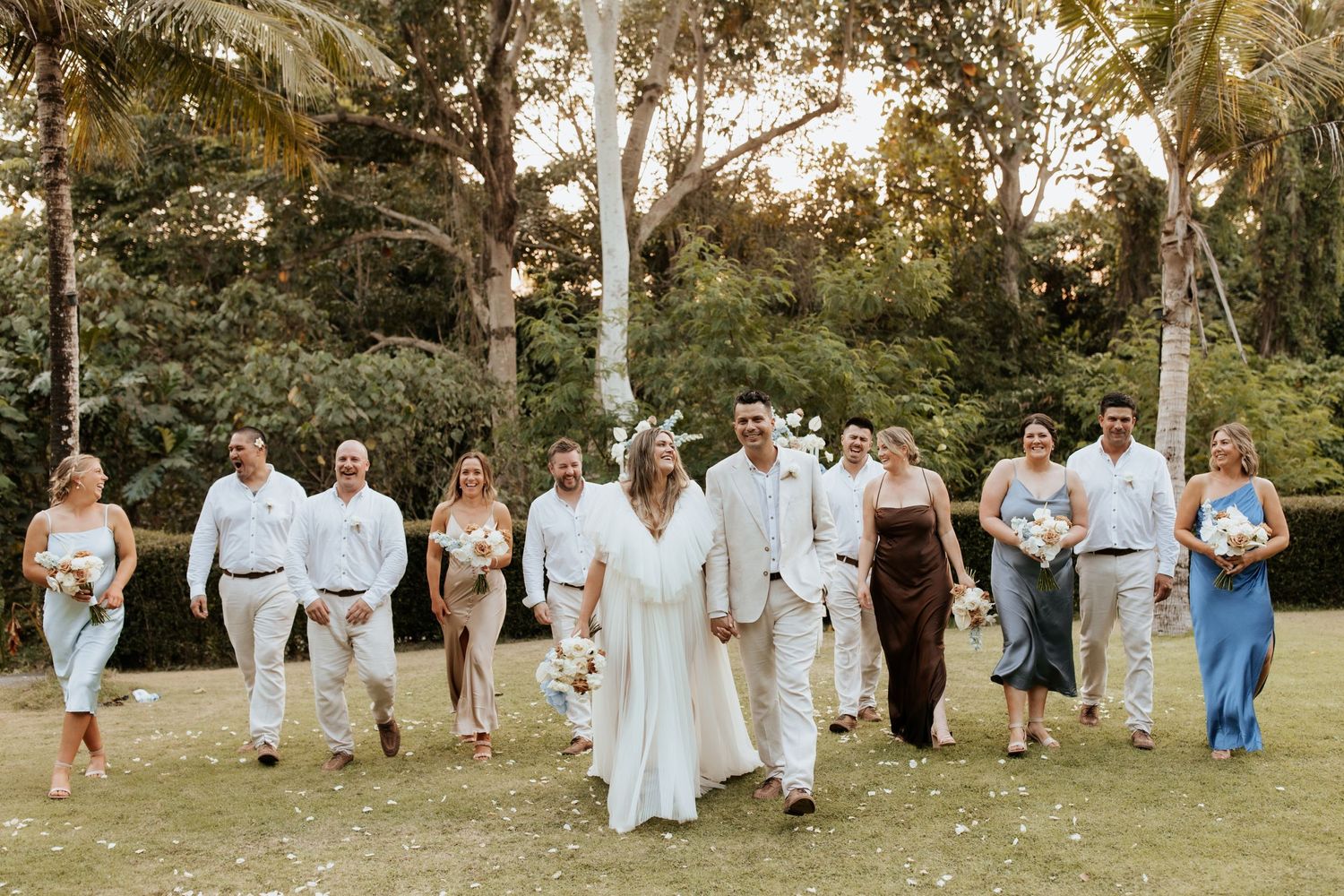 A wedding party in white and dark attire walks together through a scenic garden setting.