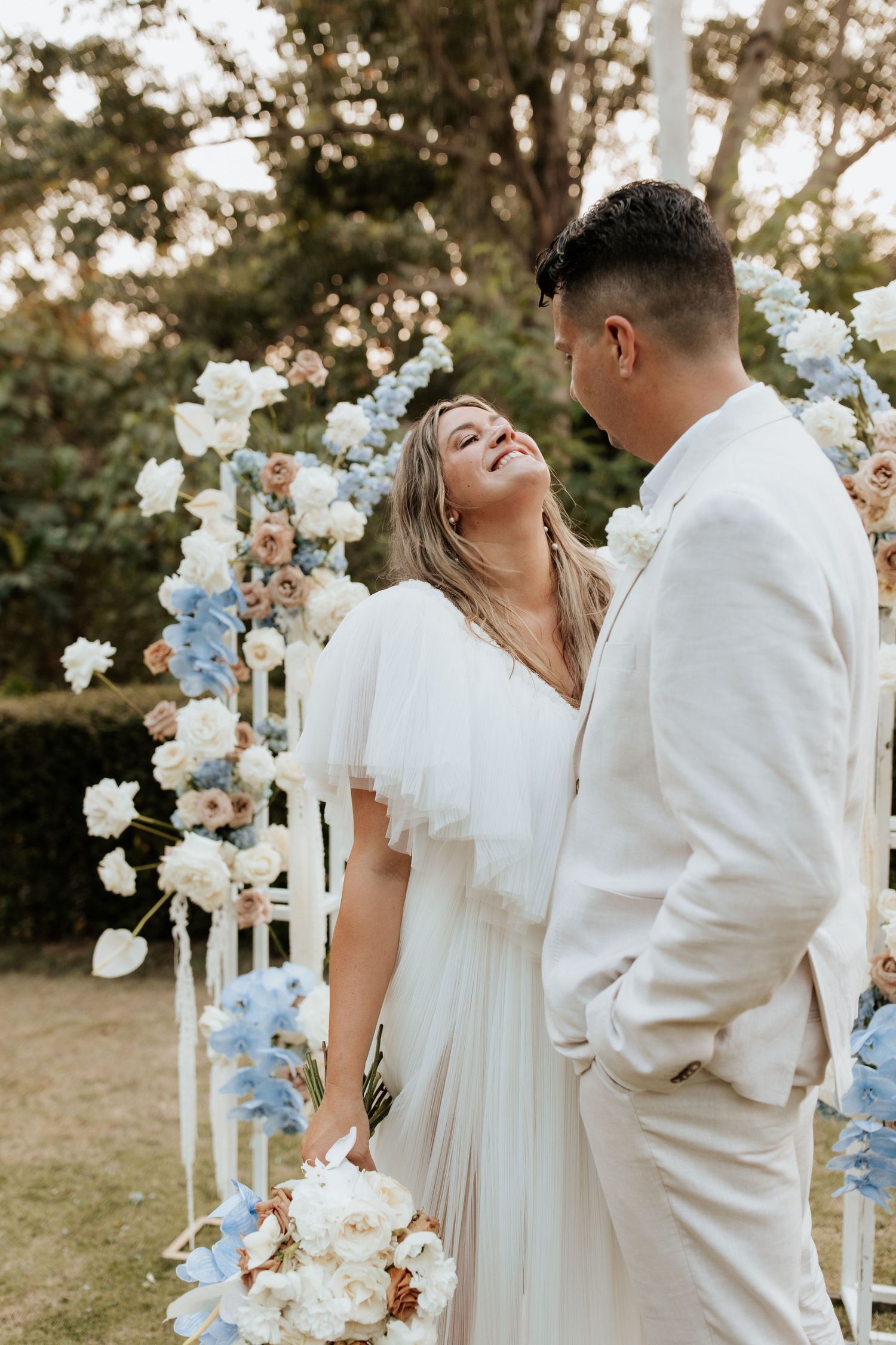 Couple shares a romantic moment in front of floral wedding arch with blue and white flowers.
