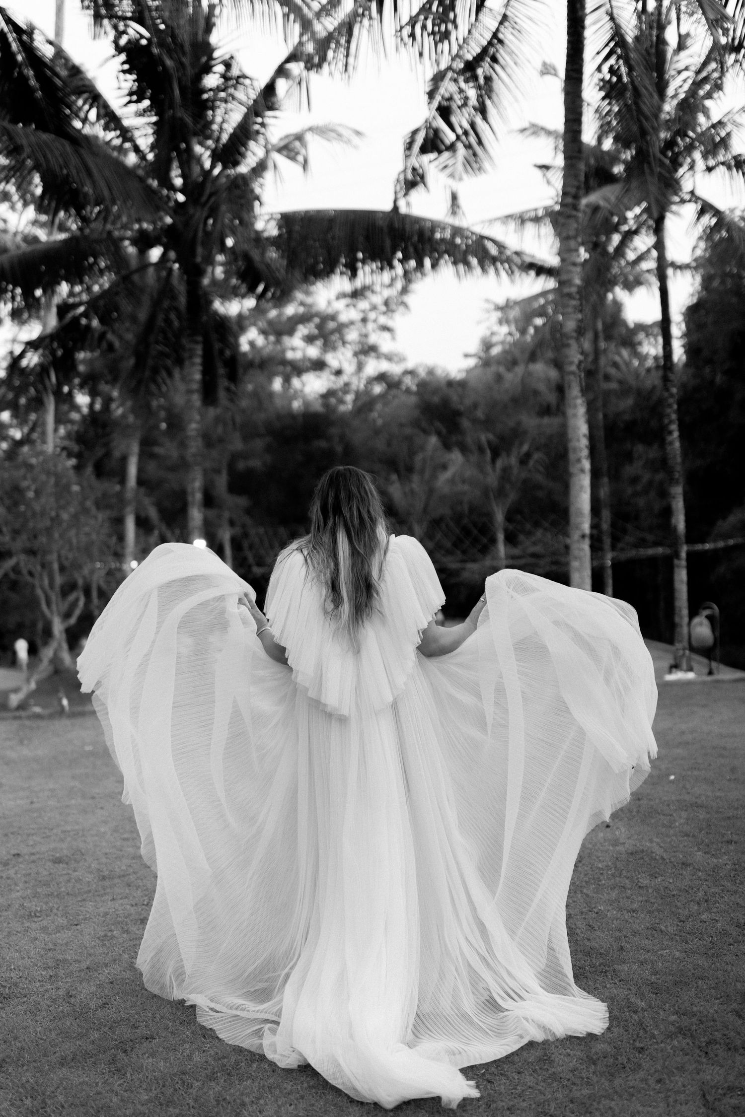 A flowing white wedding dress billows in the breeze among tropical palm trees in a black and white photograph.