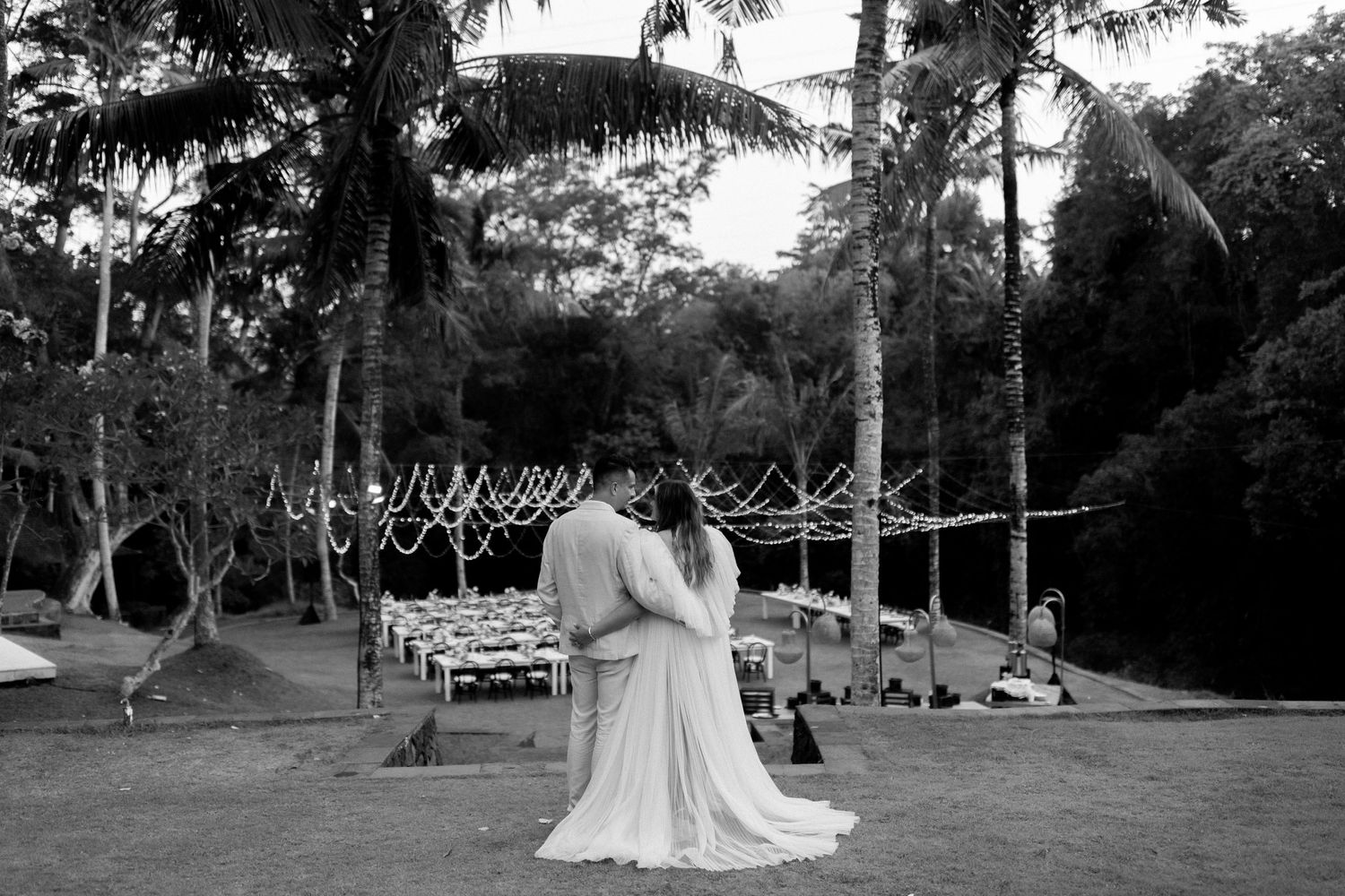 Black and white photo of a couple embracing outdoors at a rustic garden venue with string lights and trees.