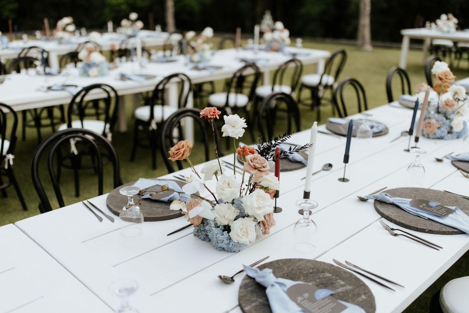 Elegant outdoor wedding reception tables set with white linens, floral centerpieces and bentwood chairs on a lawn.