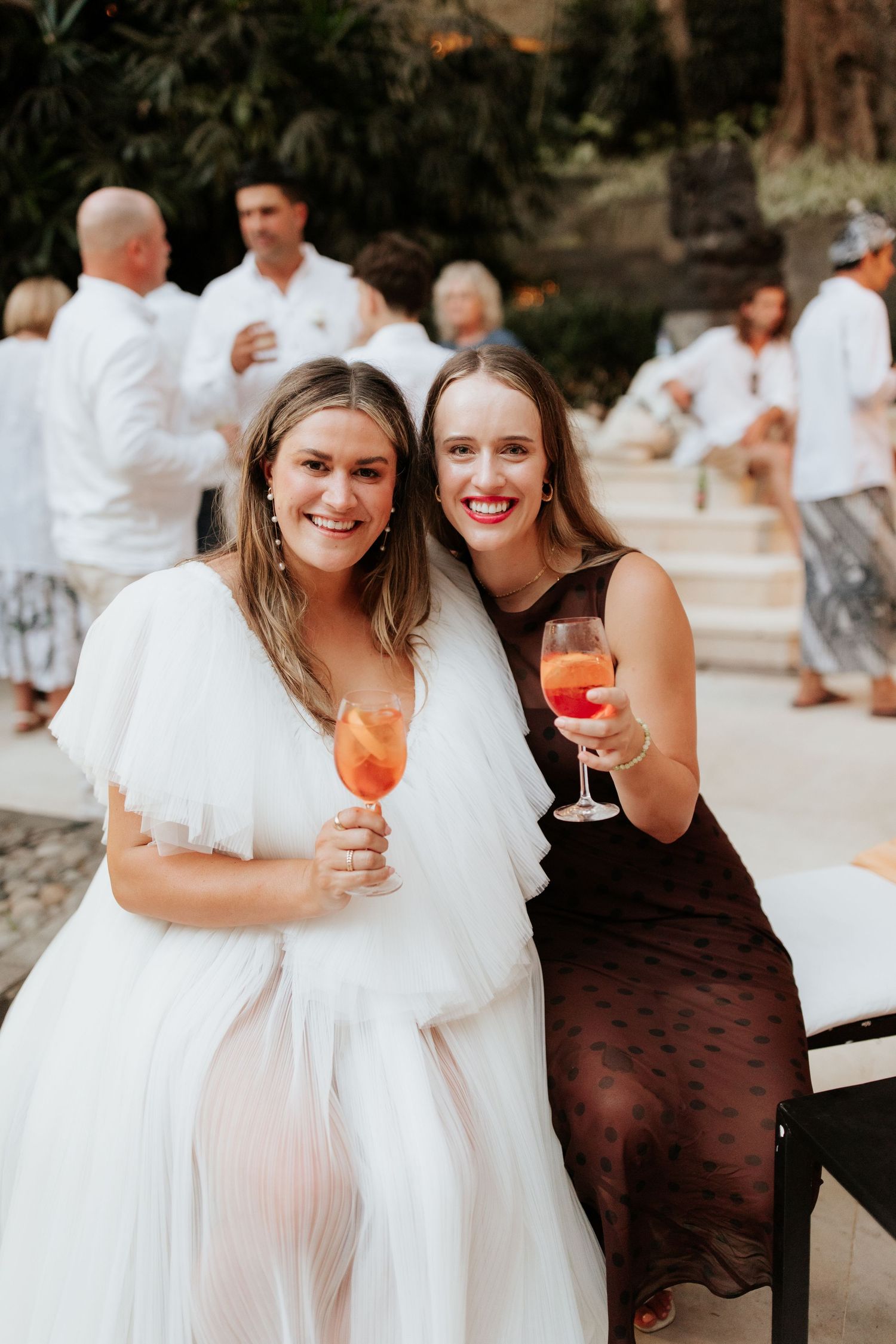 Two friends in formal attire enjoy colorful cocktails at an outdoor celebration.