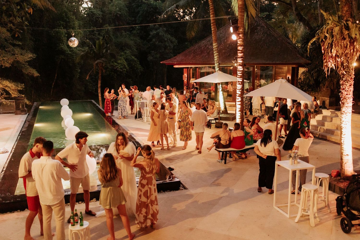 Wedding guests gather at an outdoor evening reception with string lights and palm trees creating a warm ambiance.