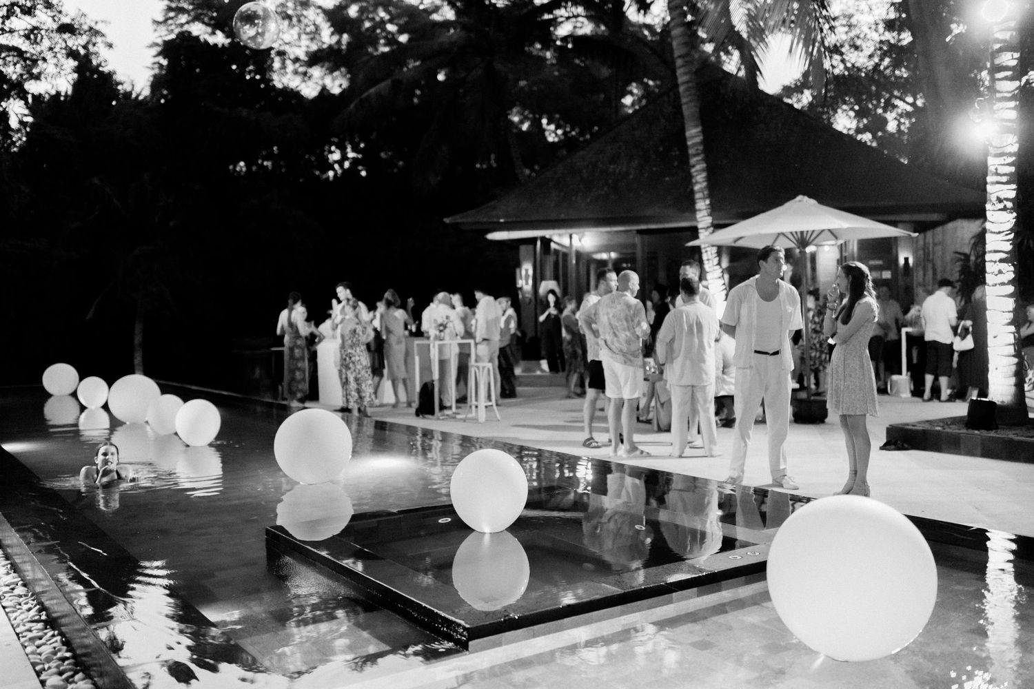Elegant nighttime poolside party with glowing balloon decorations and guests gathered around an illuminated swimming pool.