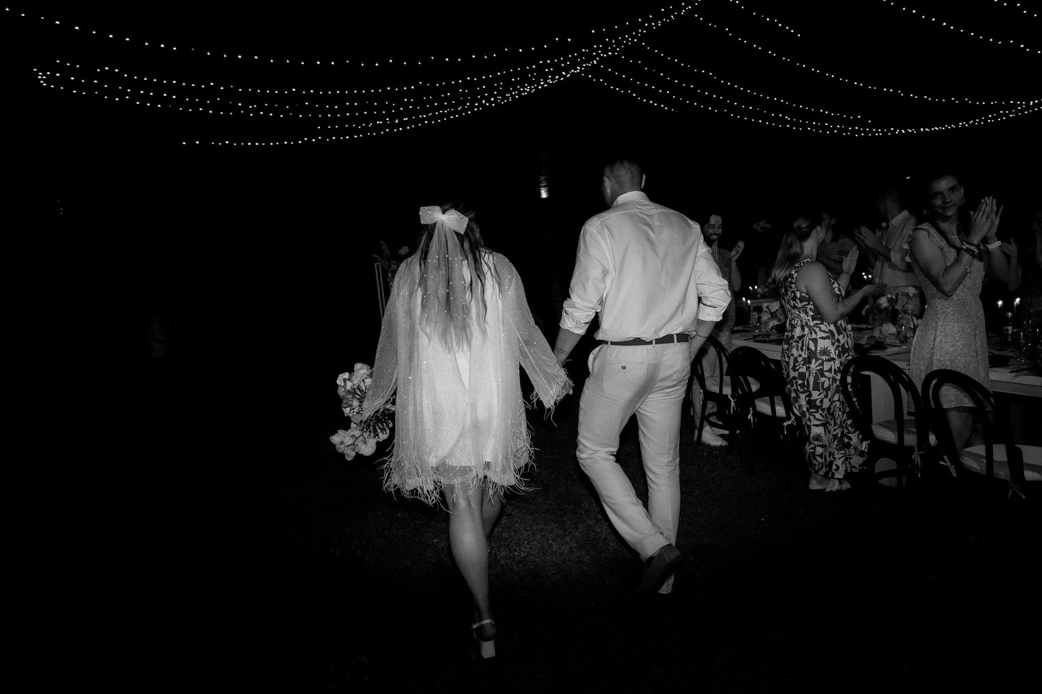 Wedding couple walking together under string lights during their night time celebration.