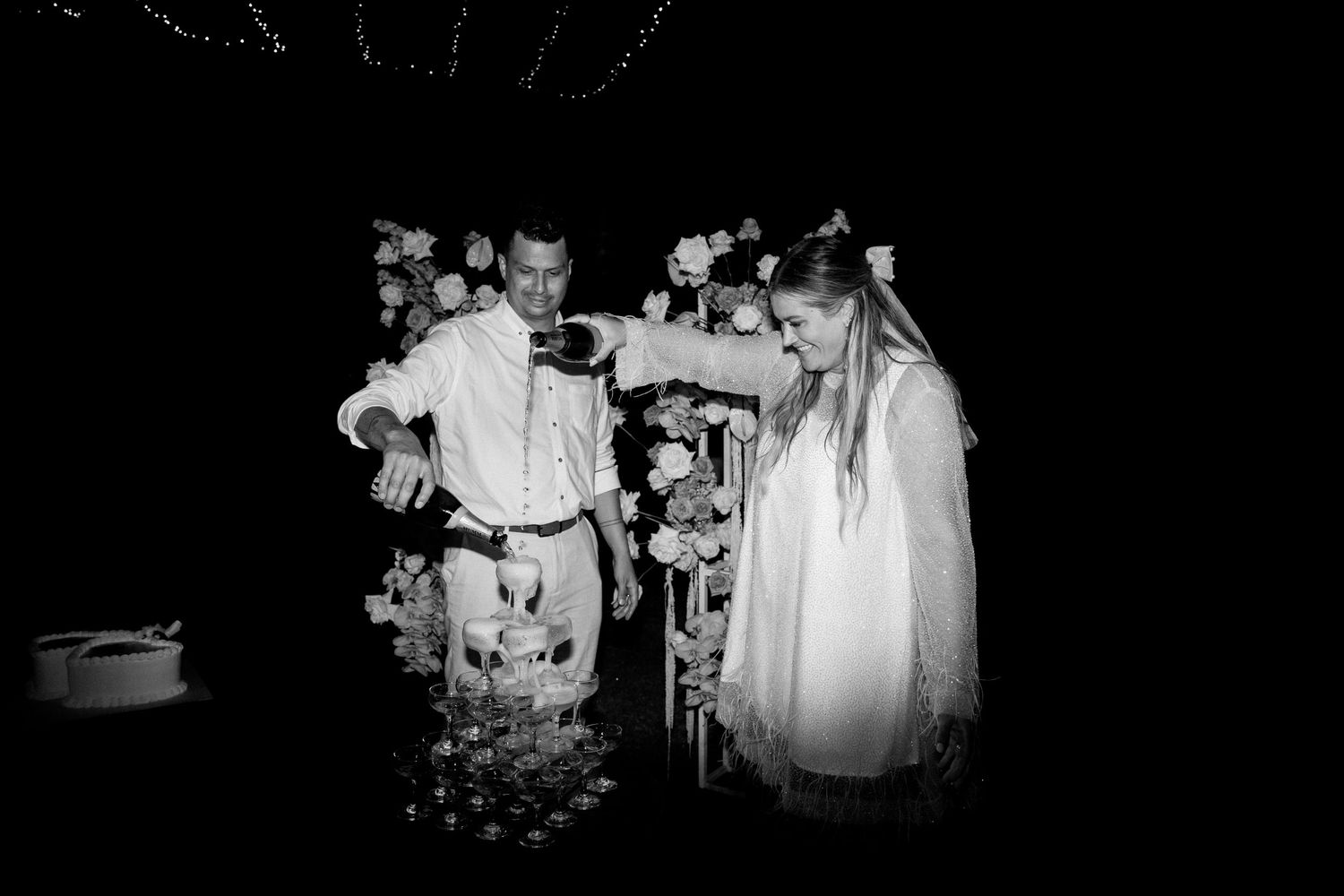 Wedding couple pouring champagne into a tower of glasses at night in black and white photography.
