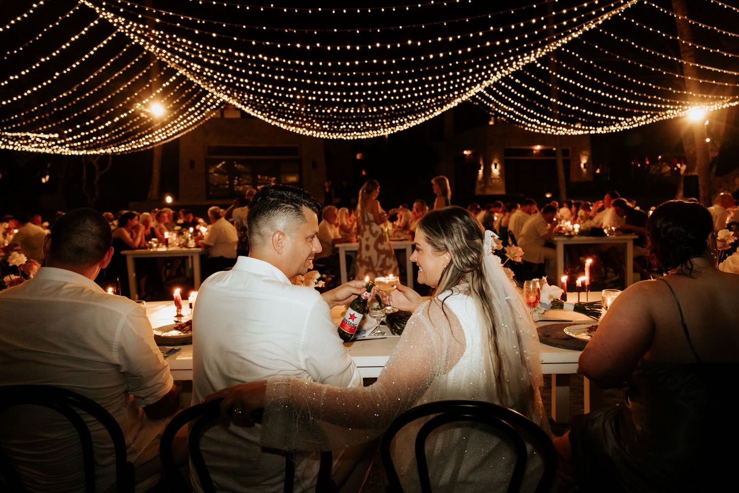 Wedding reception under glowing string lights with guests seated at tables in an outdoor venue.