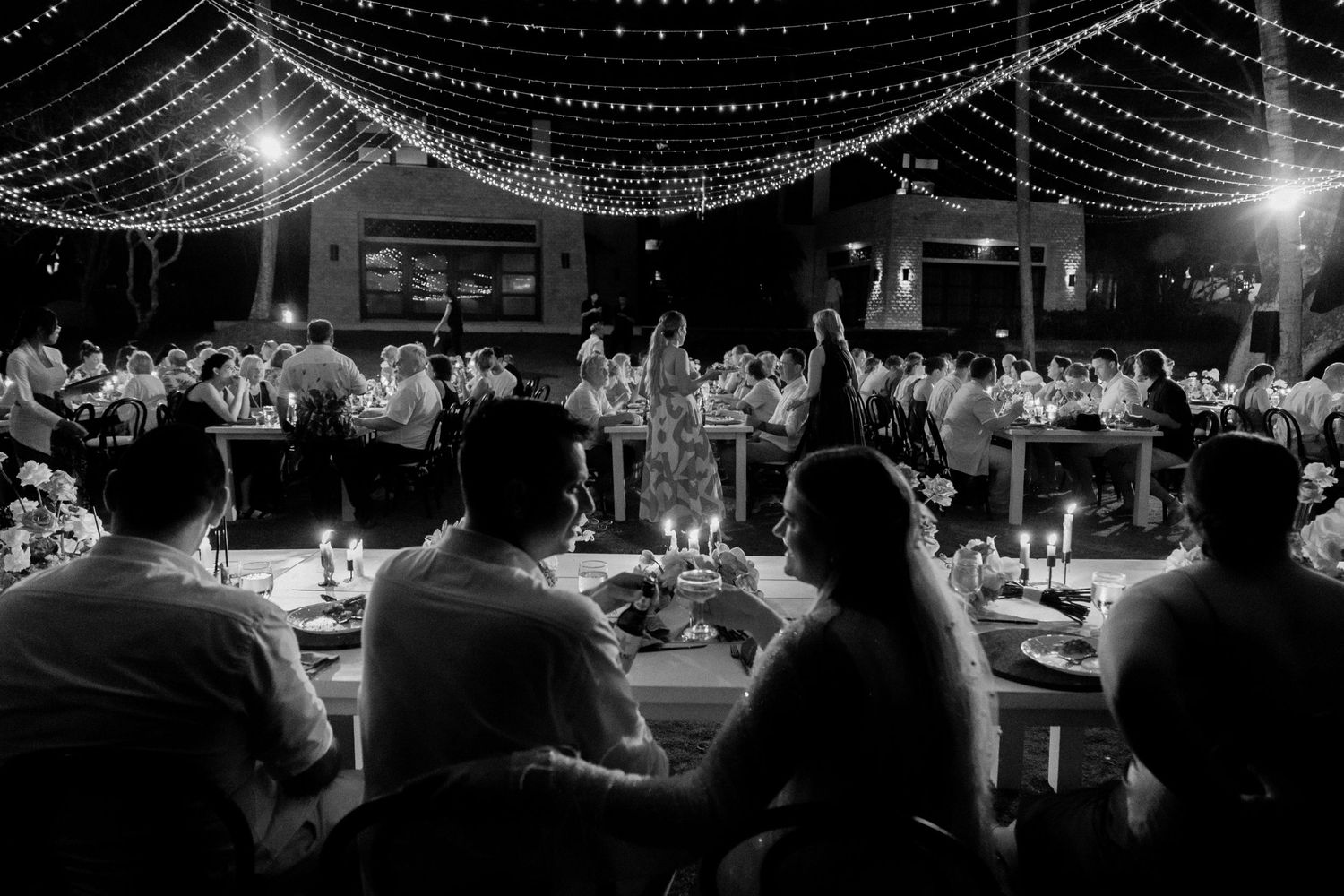 Black and white photo of wedding reception with string lights and guests seated at decorated tables.