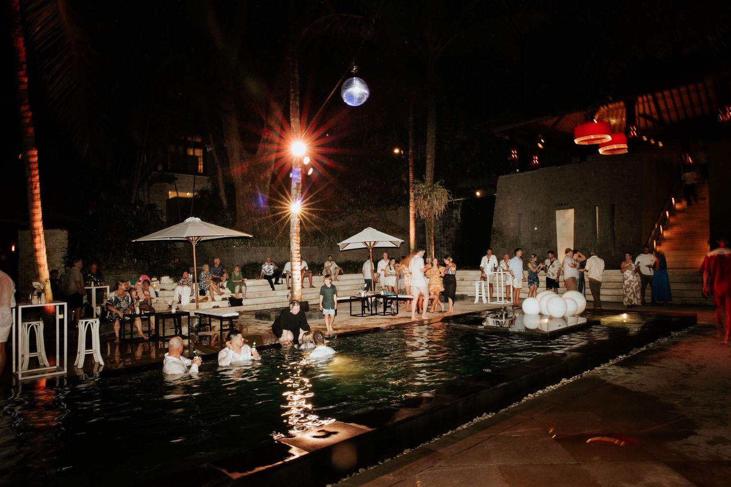 Nighttime poolside party with white umbrellas and glowing lights reflected in illuminated swimming pool.