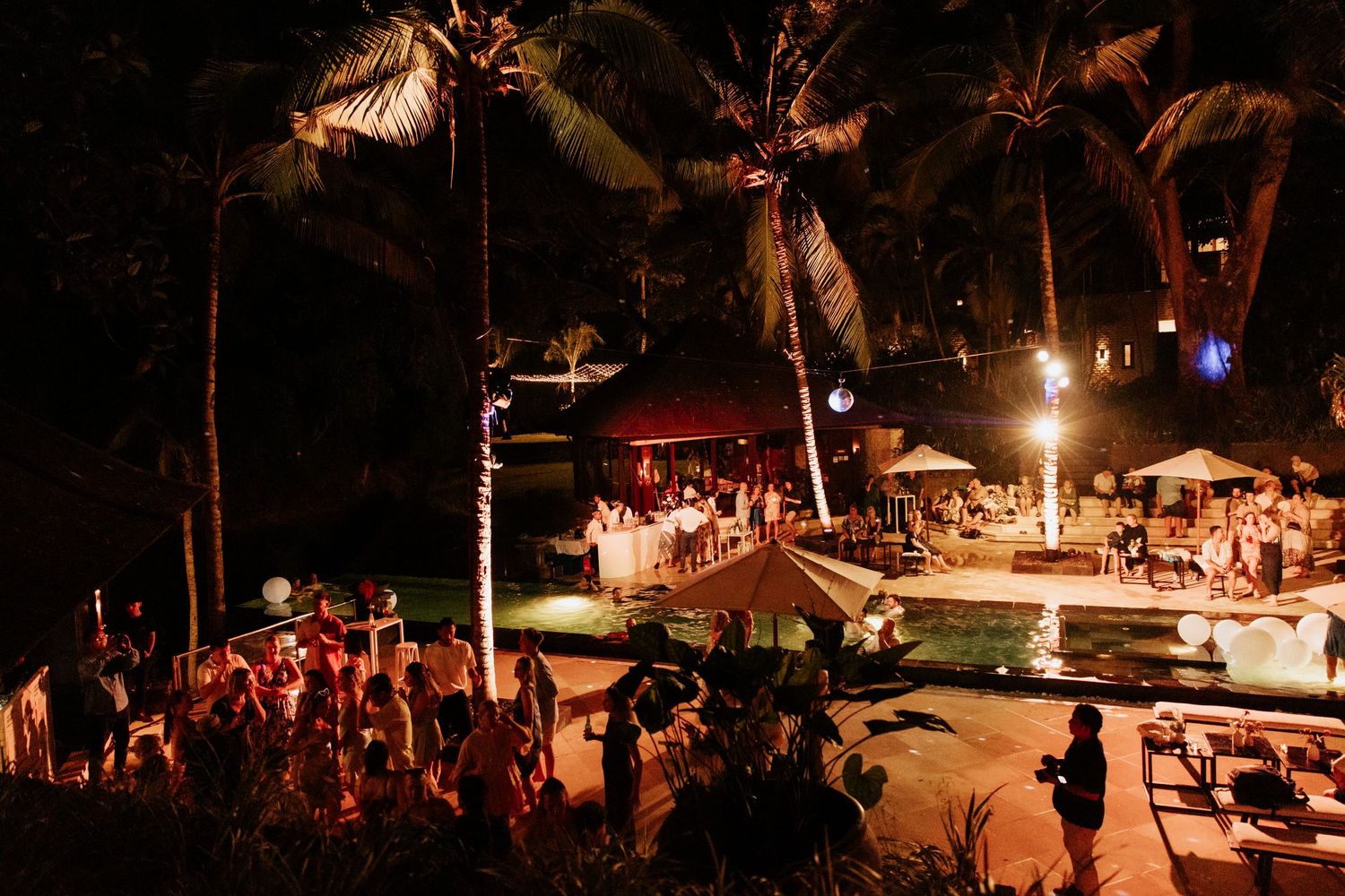 Outdoor nighttime wedding reception under palm trees with ambient lighting and crowd of guests.