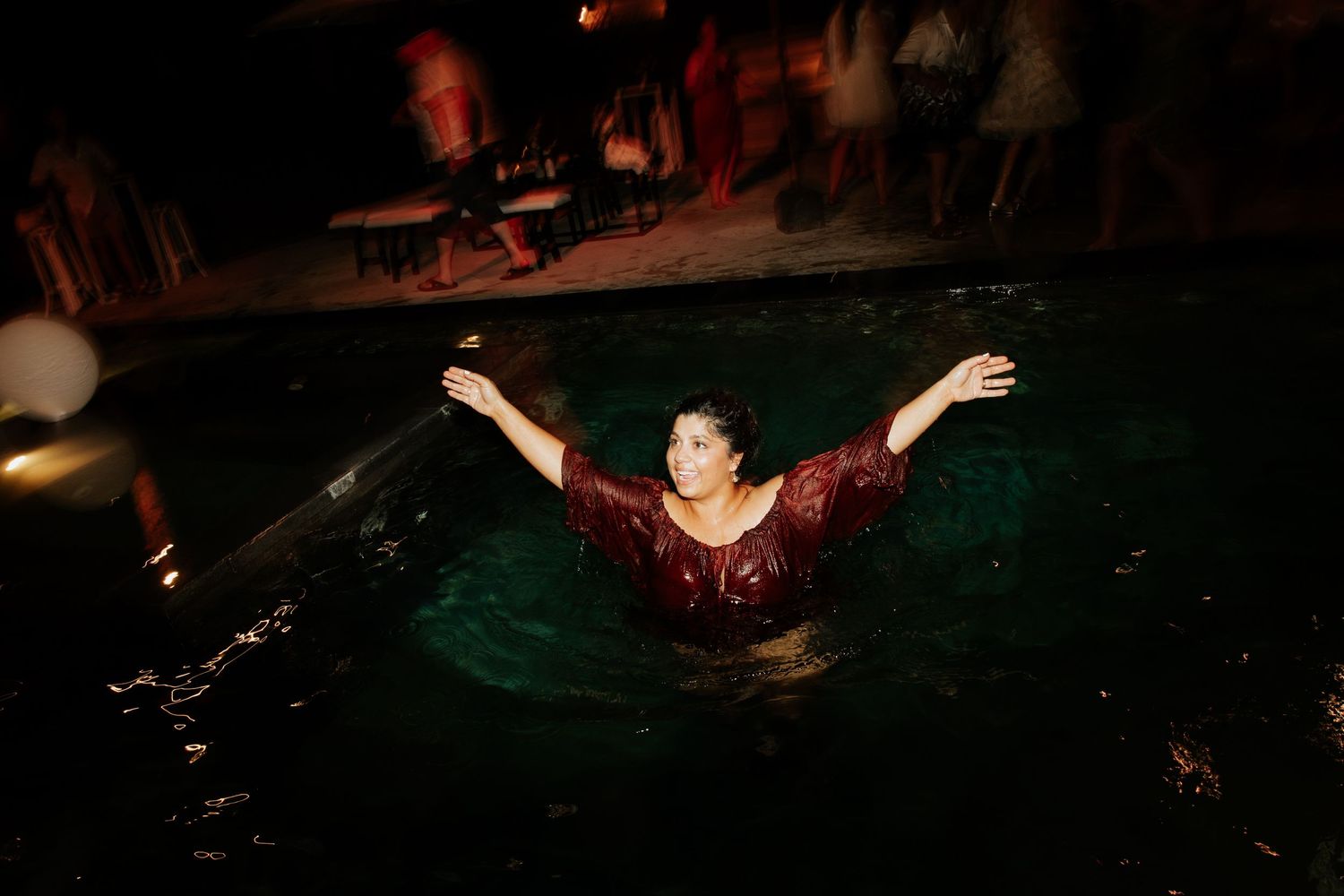 A figure emerges from a dark pool with raised arms wearing a burgundy dress in moody lighting.