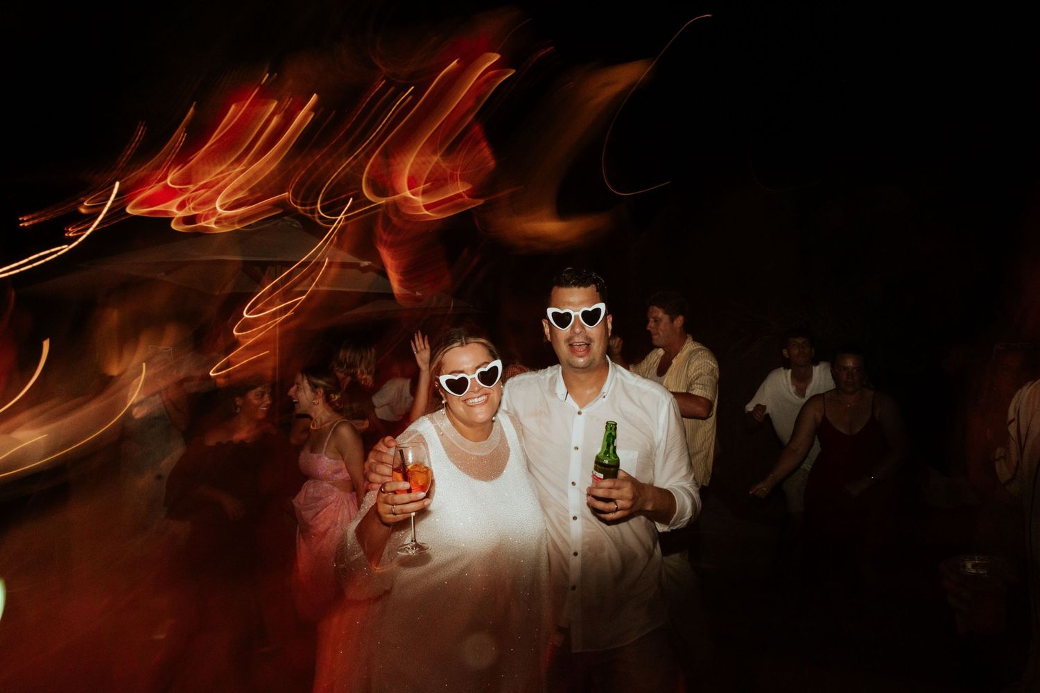 Party guests in white outfits dance under neon lights at a nighttime celebration.