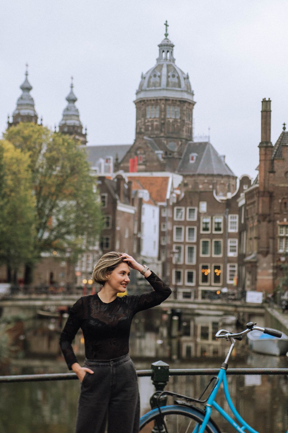 Woman with bicycle posing near historic Amsterdam canal and architecture.