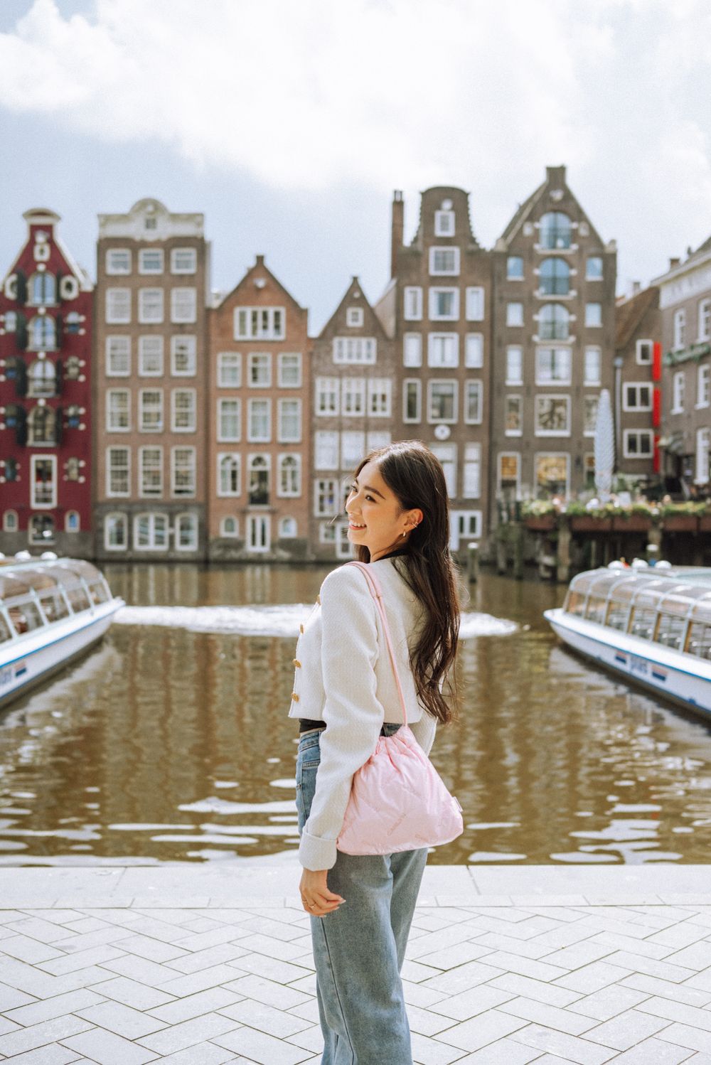 Woman in casual outfit poses against backdrop of traditional Dutch canal houses.