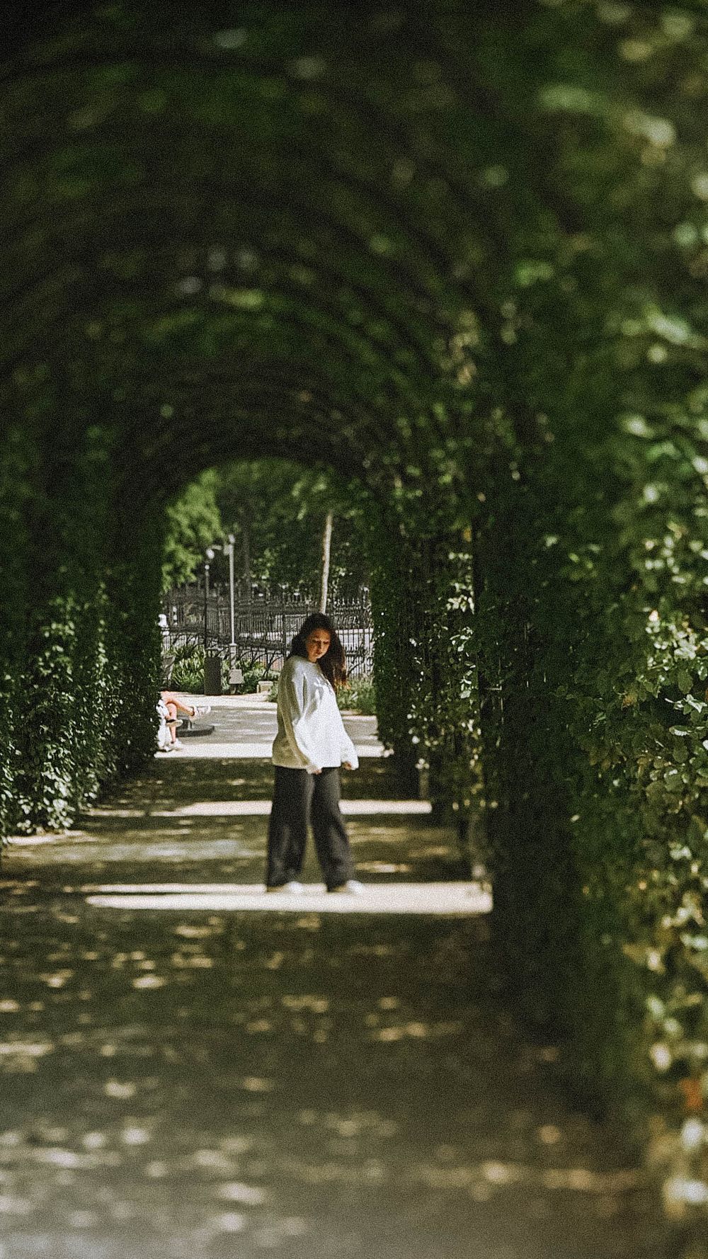 Figure walking through green garden archway in dappled sunlight.