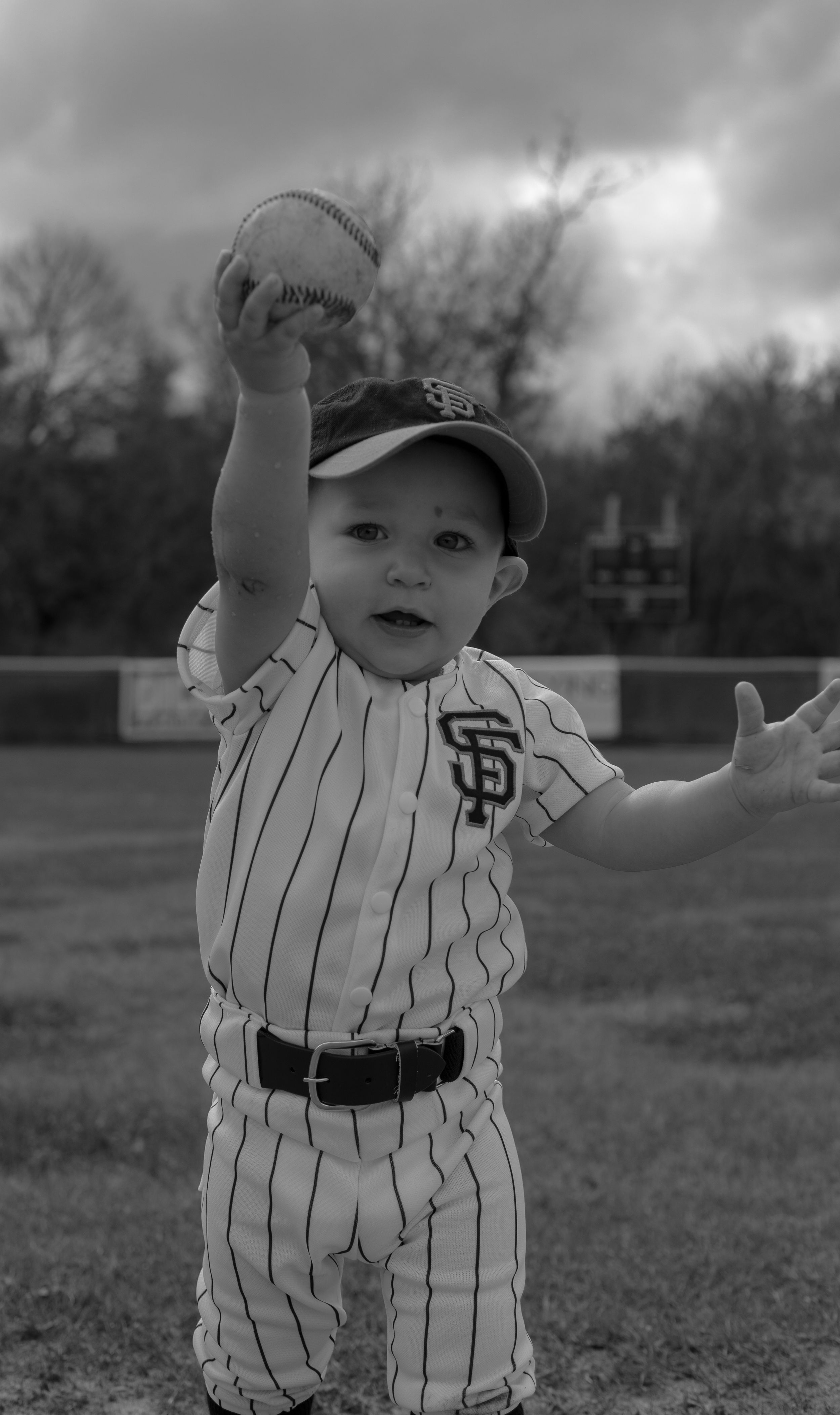 Classic black and white image, baby in vintage-style striped baseball uniform lifting baseball.