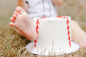 Playful cake smash setup, white baseball-themed cake near baby’s bare toes.