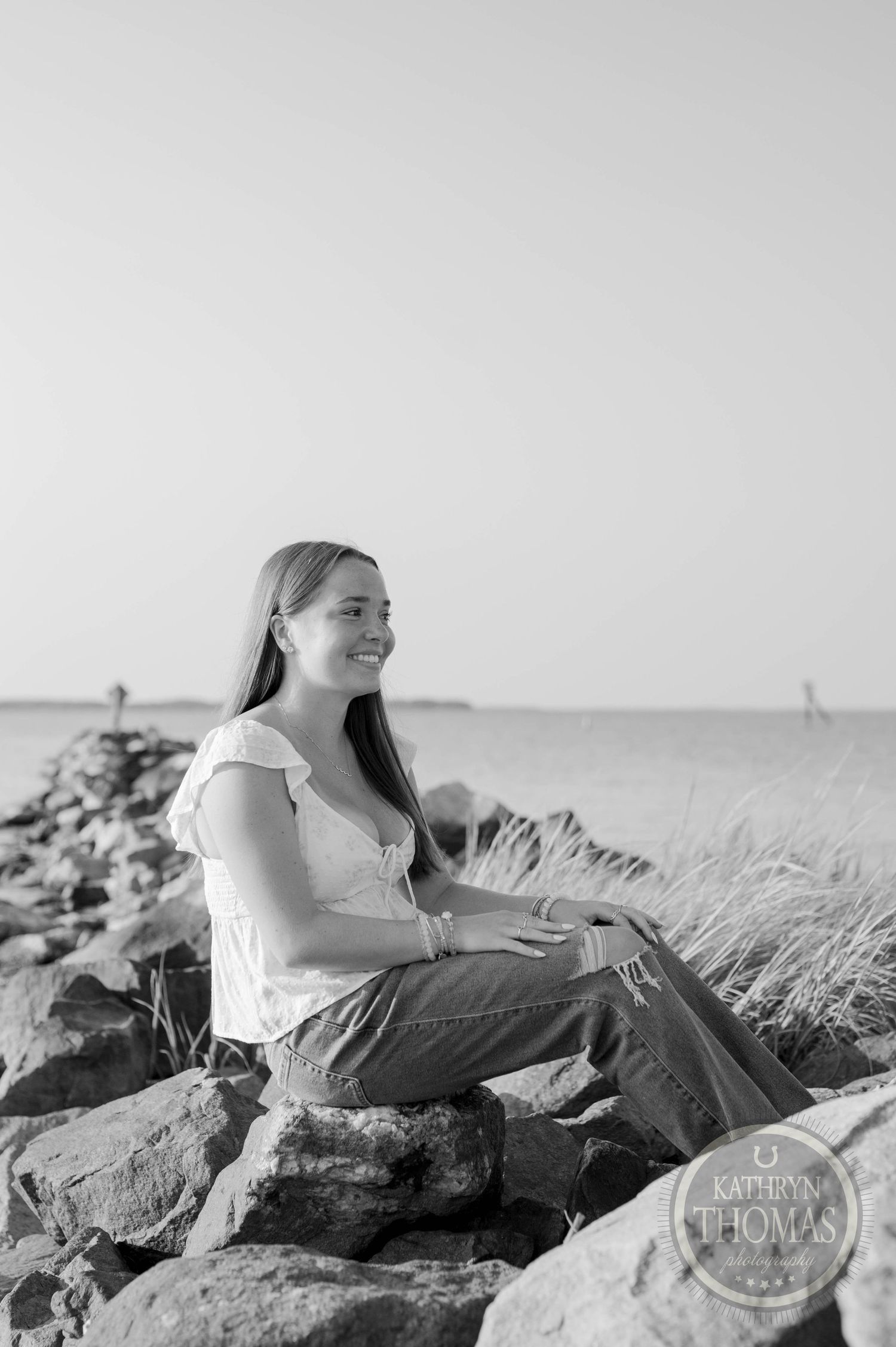 A black and white photo of a person sitting on coastal rocks at sunset wearing a white top and jeans.