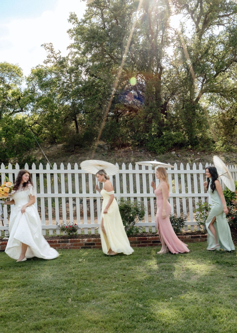 Bride in white gown walks with bridesmaids in pastel dresses on green lawn by white picket fence.
