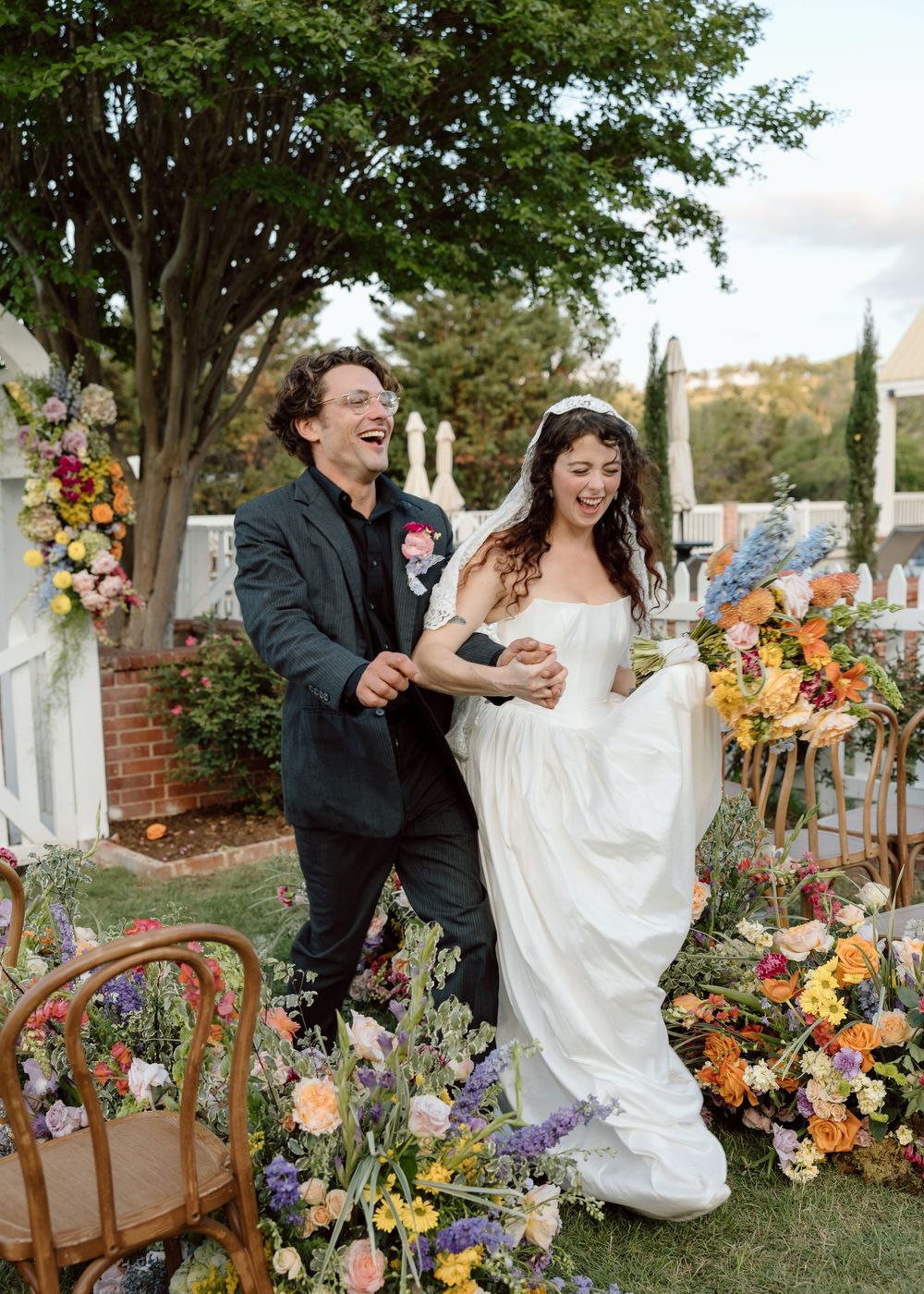 Joyful bride and groom walk through colorful floral arrangements at outdoor wedding ceremony.