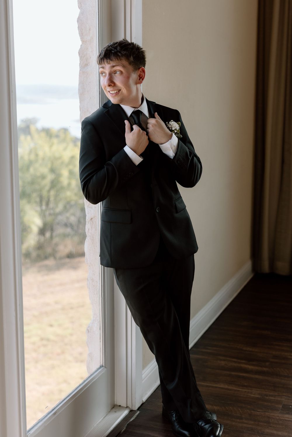 A groom in a classic black tuxedo and bowtie adjusts his jacket while standing by a window with scenic views.