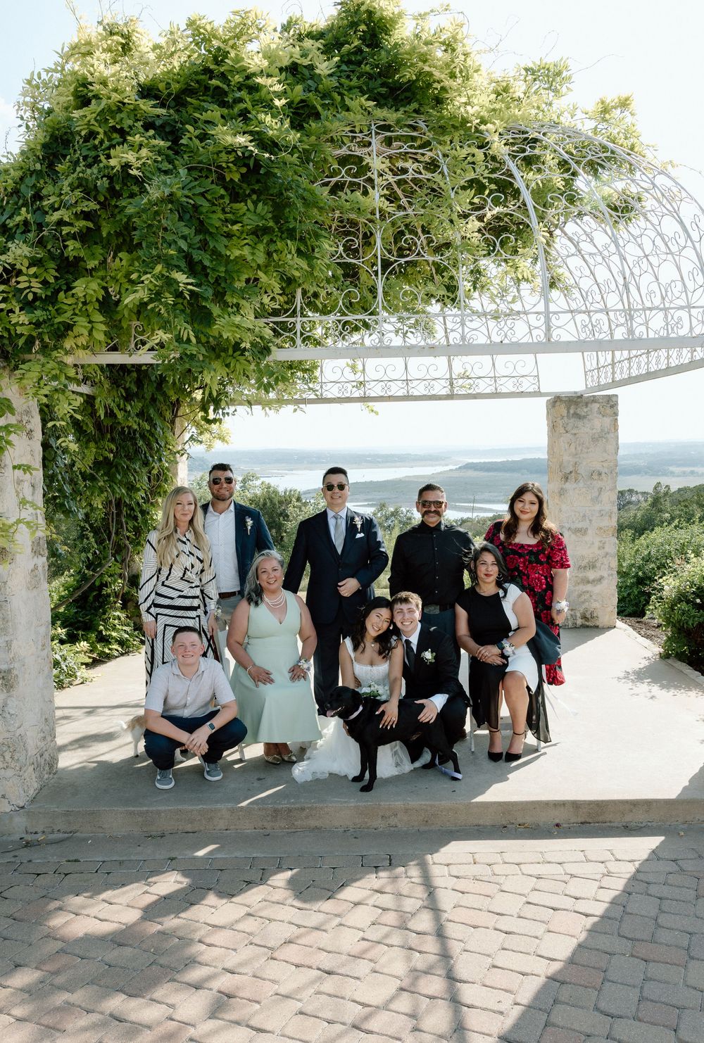 Wedding party poses under a lush greenery arch on a stone terrace with scenic views in the background.