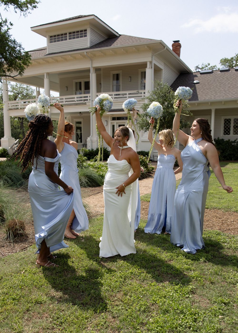 Bride in white gown celebrates with bridesmaids in light blue dresses, raising bouquets outside elegant Southern home.