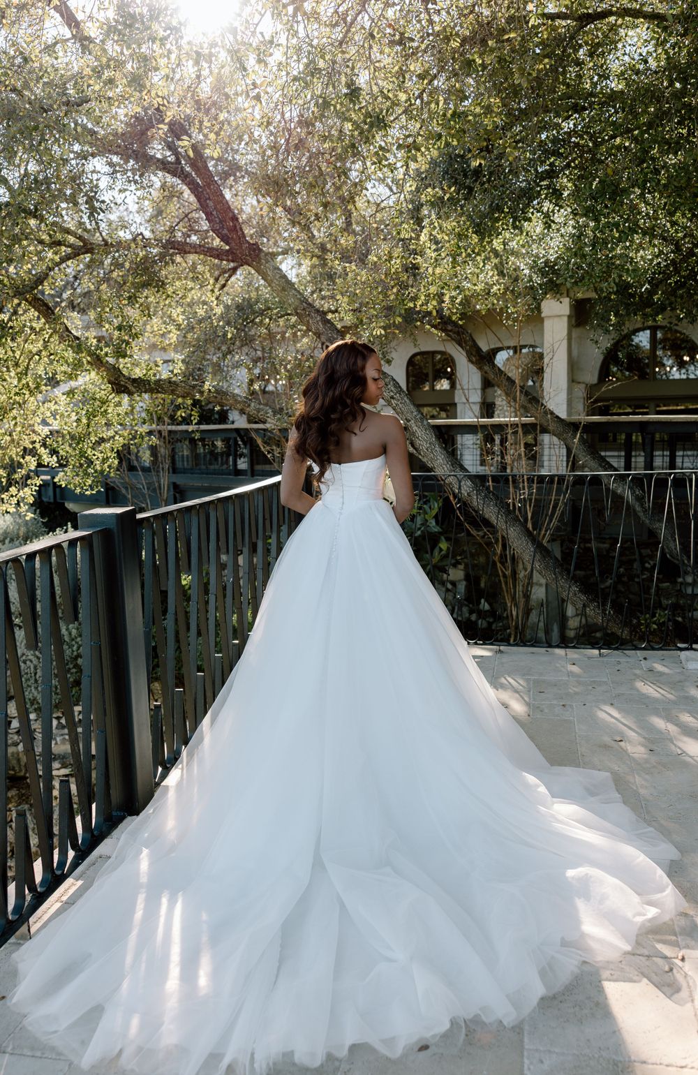 Elegant white wedding gown with full tulle skirt and long train photographed from behind on a sunlit balcony.