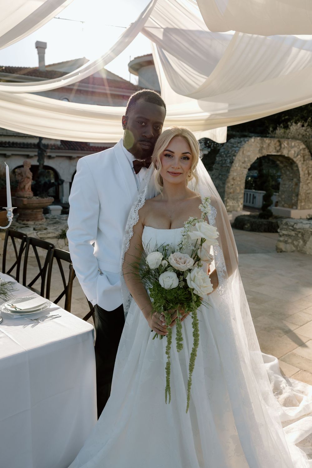 A bride in a white gown with cascading veil holds a white rose bouquet under elegant draped fabric at a stone venue.