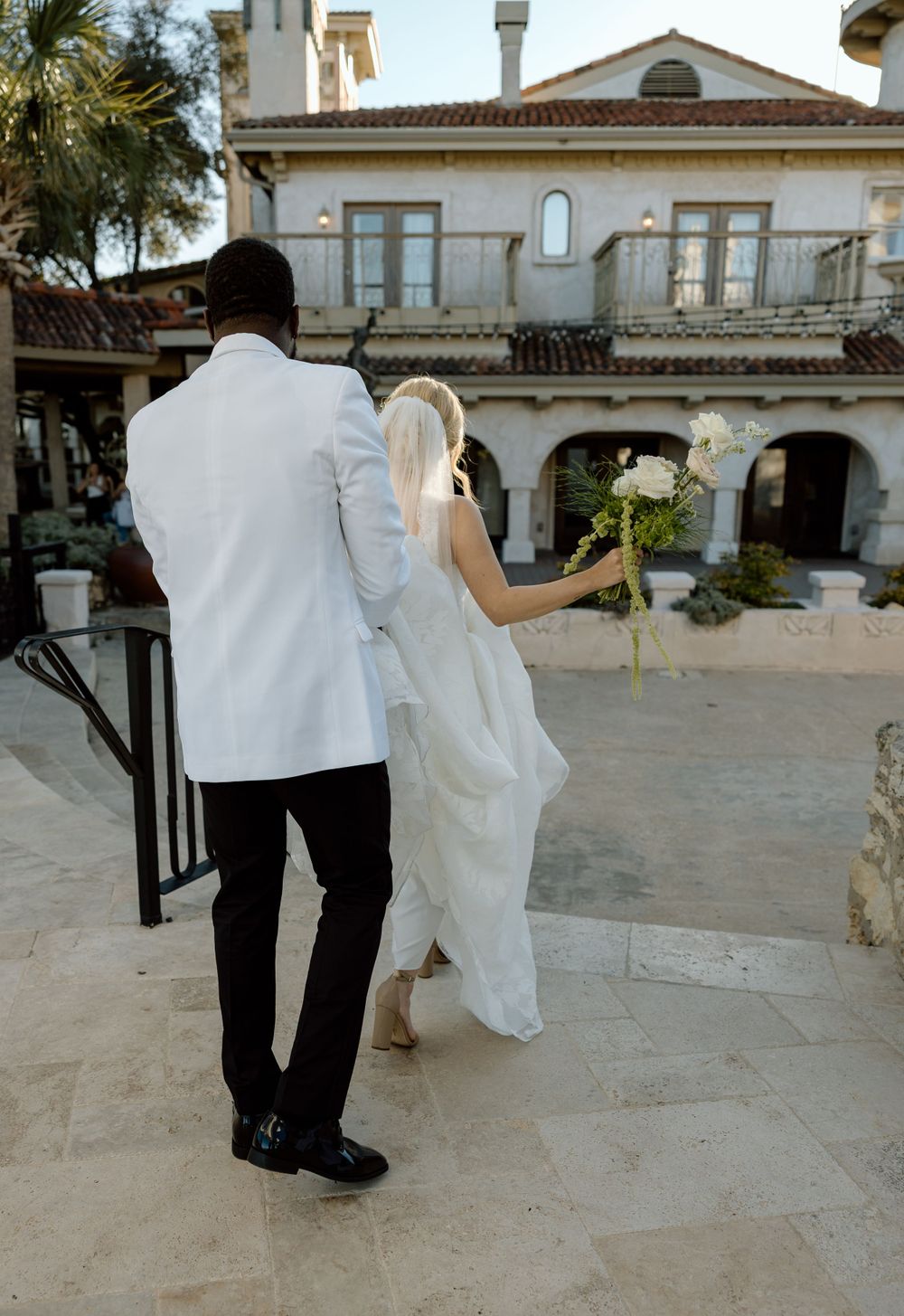A couple in wedding attire ascends stone steps outside a Mediterranean-style building with arches and palm trees.