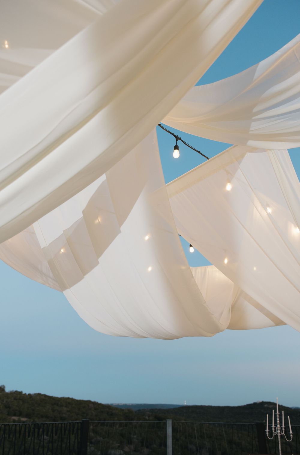 Draped white fabric and string lights create an elegant canopy against a blue sky with trees silhouetted in the distance.