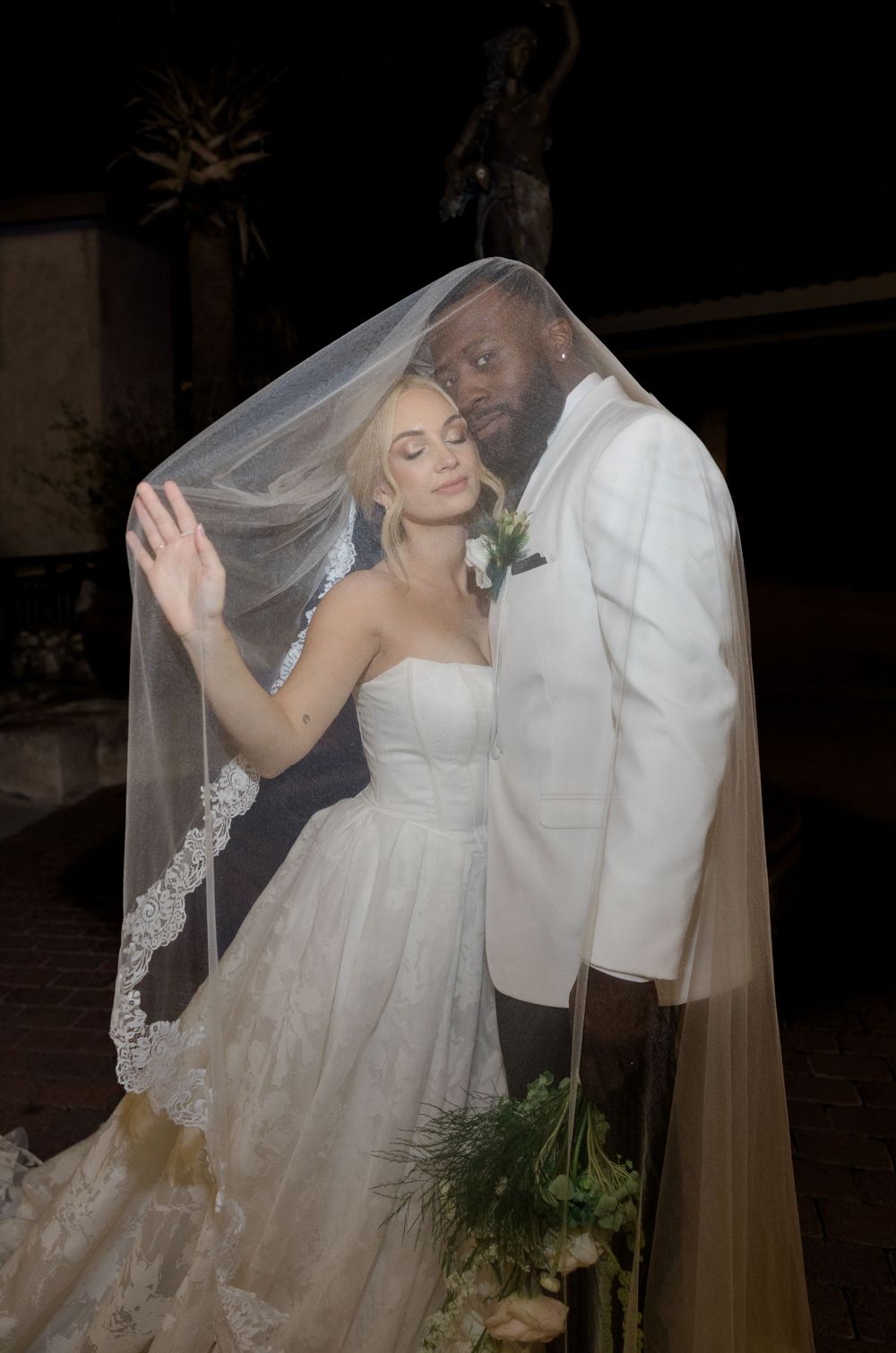 A bride and groom share an intimate moment under her flowing veil at their evening wedding ceremony.
