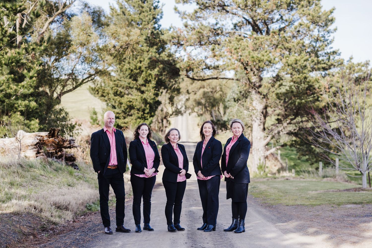 A group of people wearing black and pink formal attire stand together on a path surrounded by pine trees.