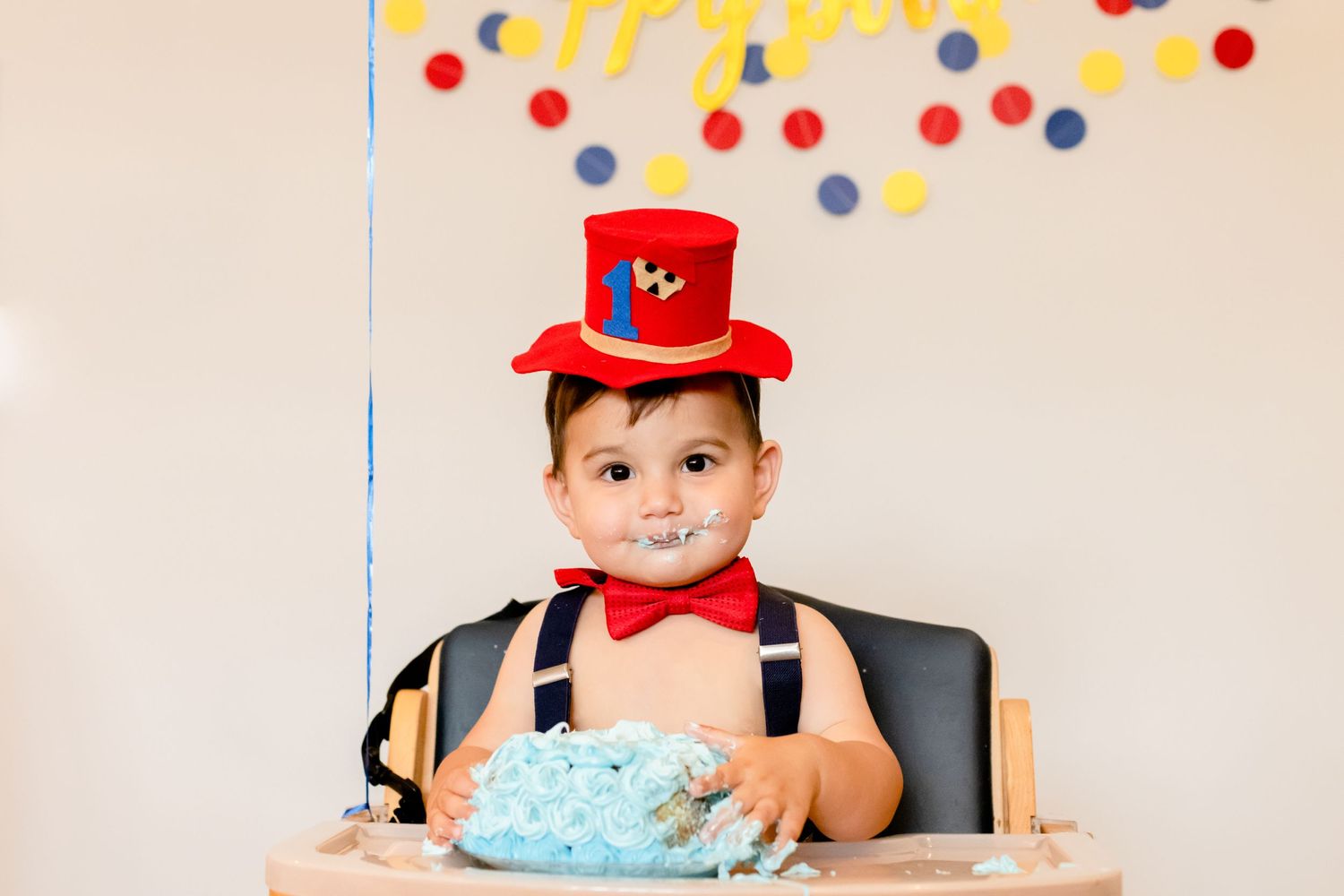 First birthday celebration scene with blue cake and red top hat amidst colorful polka dot decorations.
