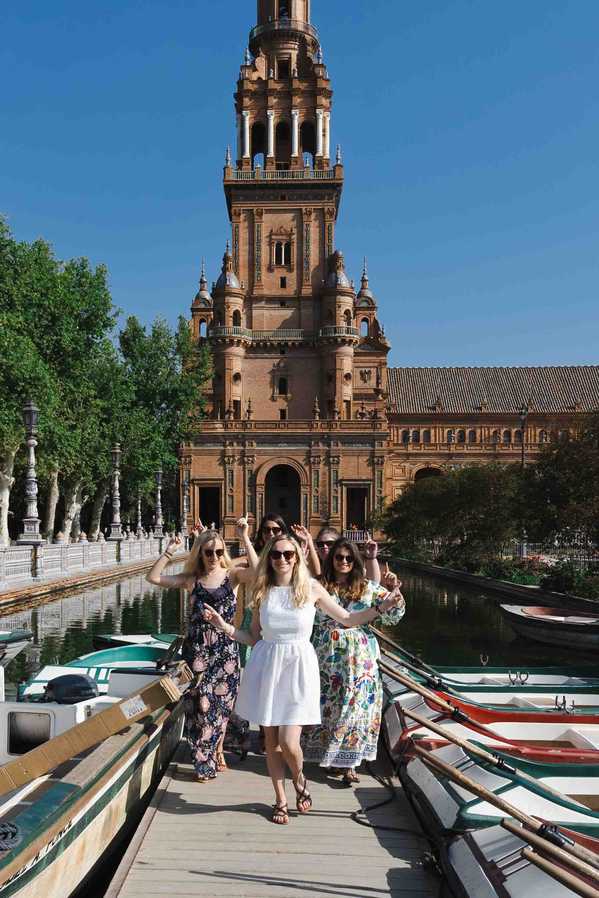 Group poses on a bridge near boats with the ornate Plaza de España tower and architecture in Seville, Spain.