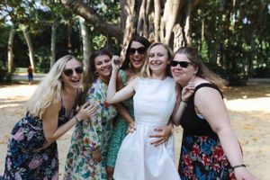 A group of friends in colorful summer dresses laugh together while posing outdoors in a wooded setting.