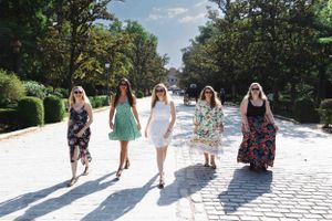 A group of five friends in sundresses walk along a bright sunlit garden path on a summer day.