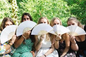 People holding white folding fans up to their faces while sitting outdoors in a garden setting.