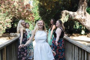 Group in floral dresses and a white dress pose together on a wooden boardwalk surrounded by lush greenery.
