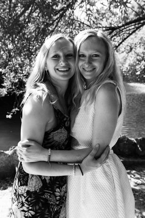 Two people in floral and polka dot dresses embrace while smiling outdoors near water on a sunny day in black and white.