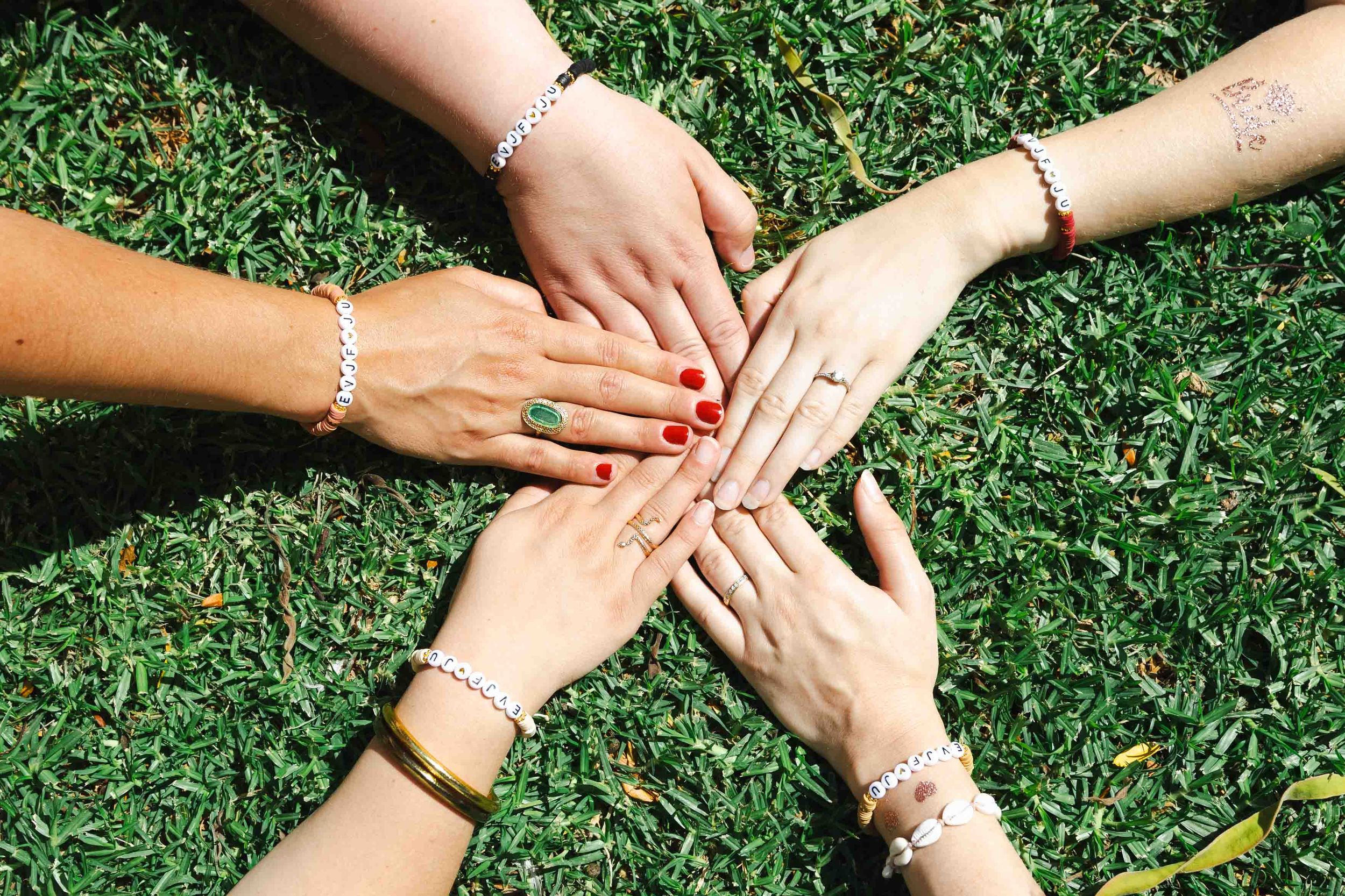 Multiple hands with jewelry and bracelets join together in a circle formation on green grass.