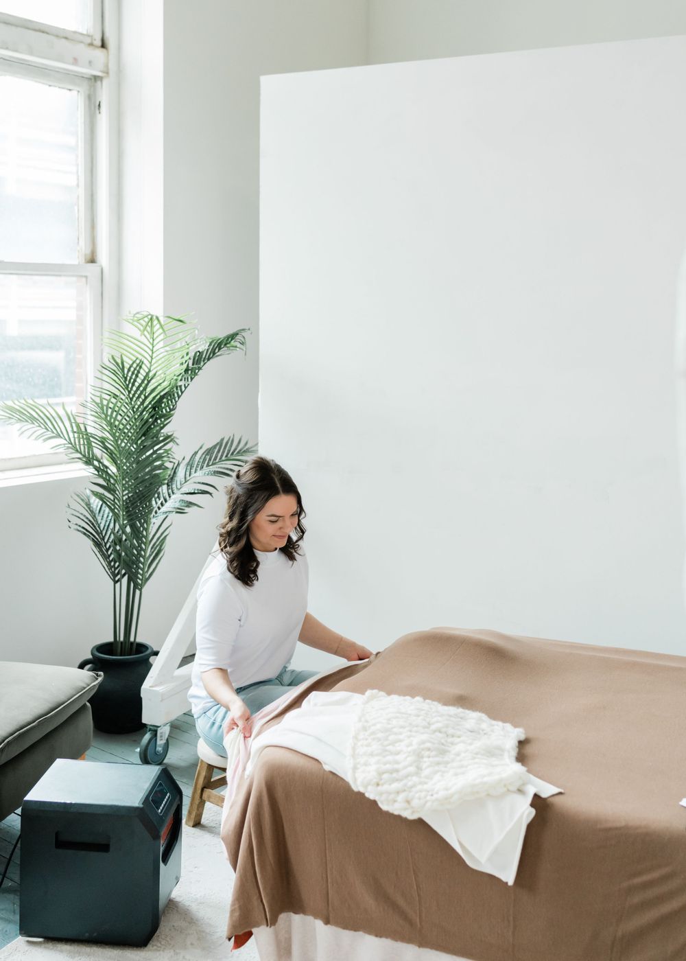 A minimalist white room features a potted palm plant and window while a person in light clothing decorates with white fabrics.