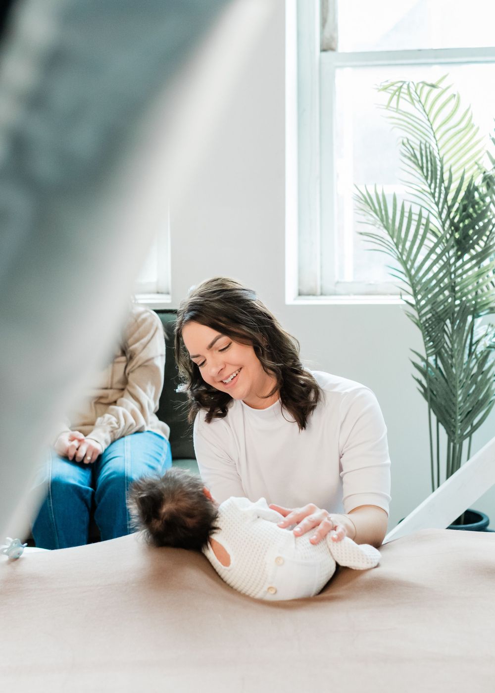 Mother and baby sharing a tender moment while relaxing on a white couch near a window with tropical plant decor.